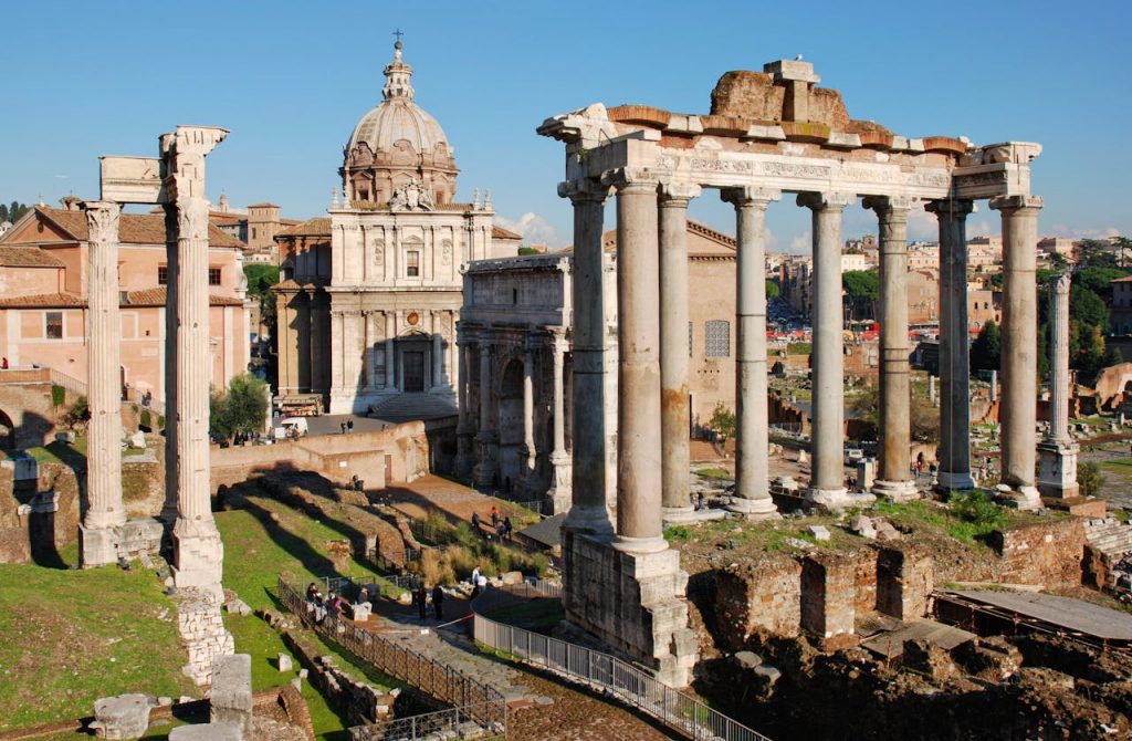 View of the Roman Forum ruins in Rome with ancient columns and historic buildings under a clear blue sky.