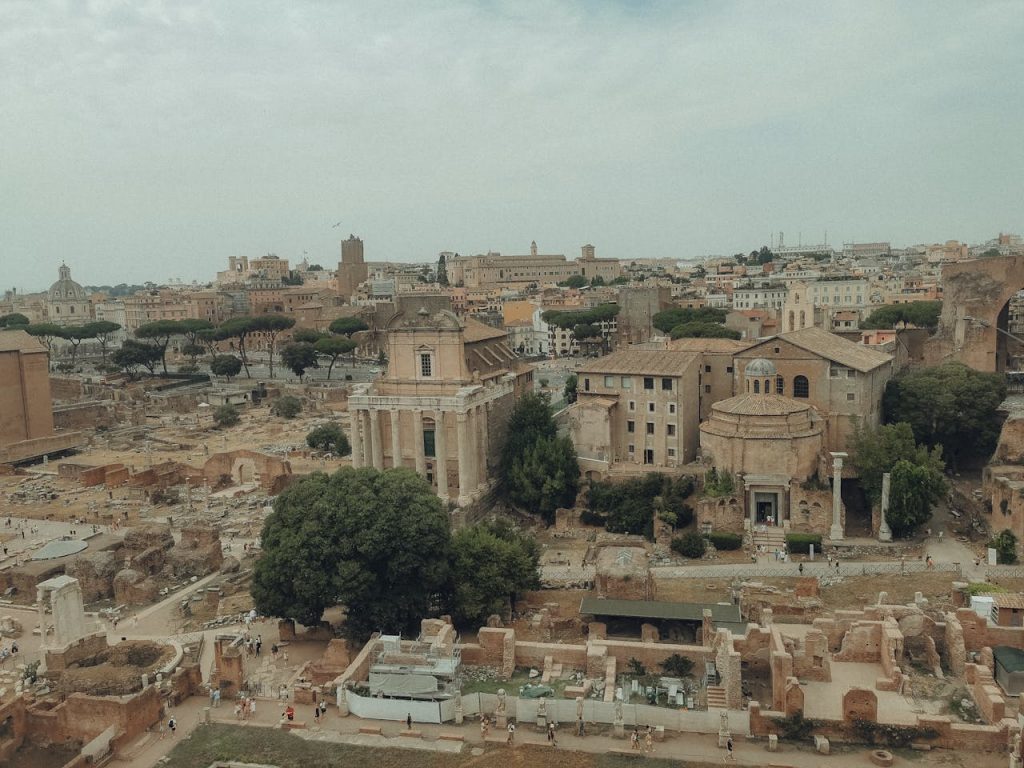 Aerial view of the Roman Forum showing ancient ruins, historic temples, and surrounding buildings under a cloudy sky in Rome, Italy.